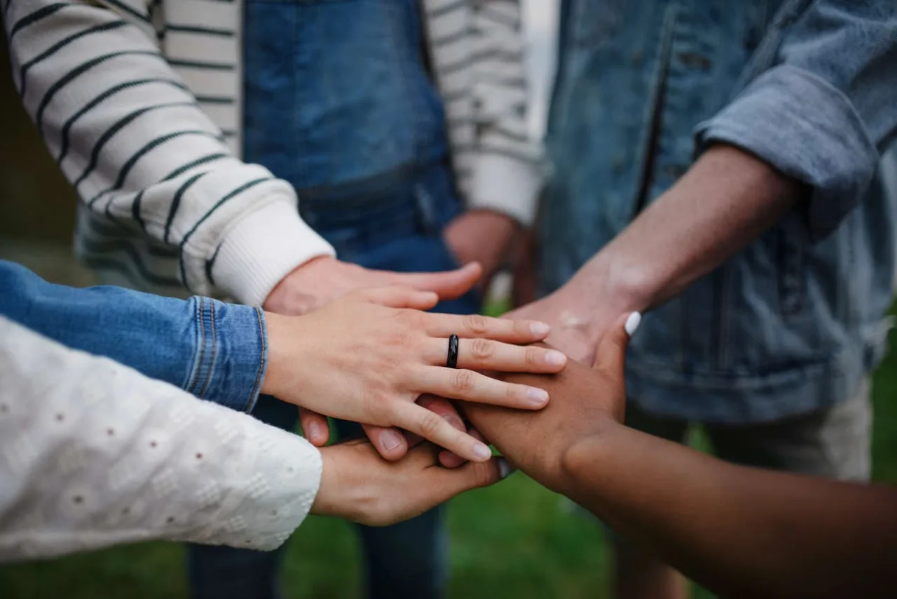 A diverse group of hands, stacked together symbolizing unity and teamwork, with one hand wearing a black ring, on a grassy background. A diverse group of hands, stacked together symbolizing unity and teamwork, with one hand wearing a black ring, on a grassy background
