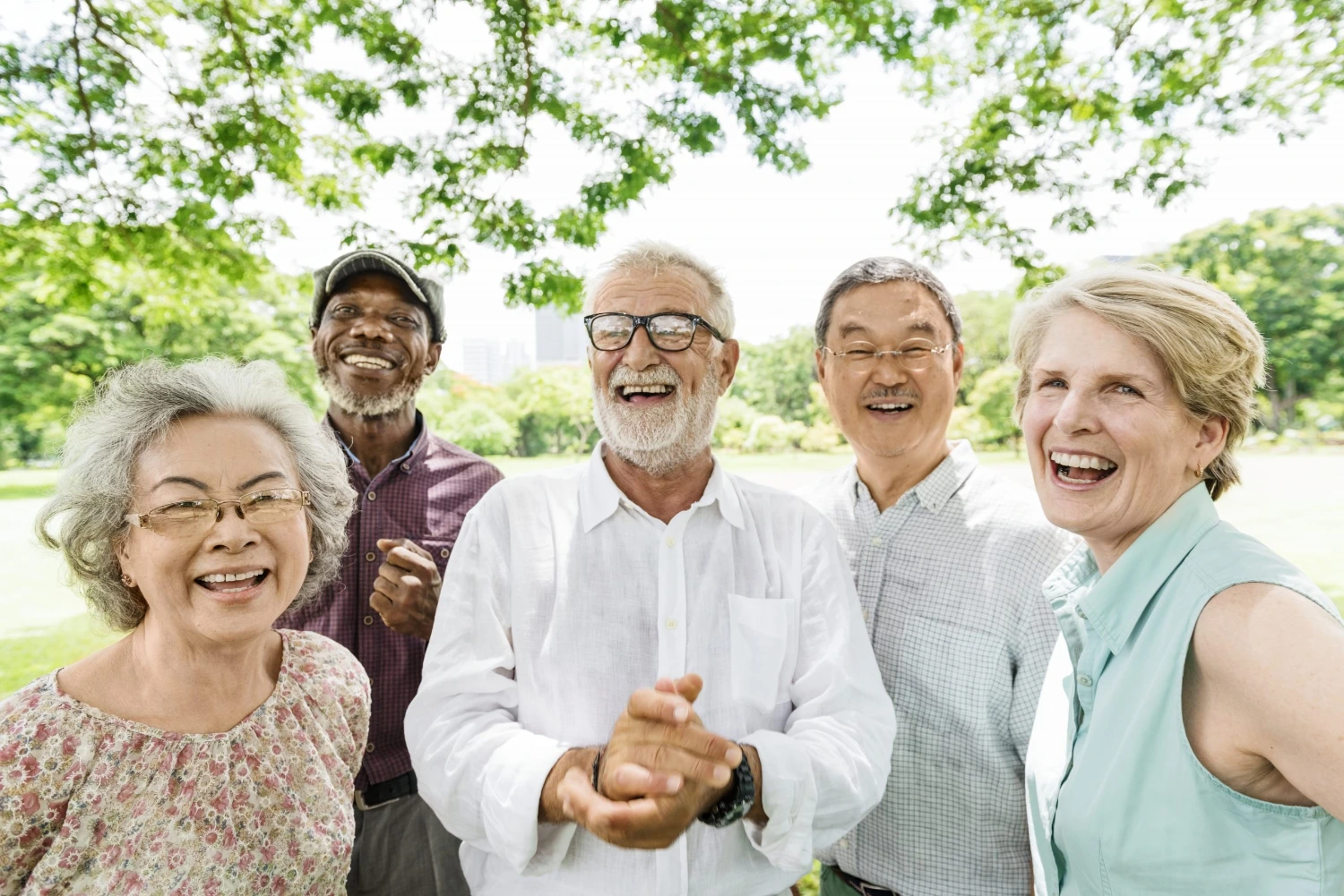 A diverse group of five adults standing outdoors under lush green trees, engaged in conversation with a vibrant park in the background. A diverse group of five adults standing outdoors under lush green trees, engaged in conversation with a vibrant park in the background